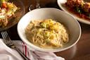 A bowl of creamy pasta topped with roasted cauliflower sits on a wooden table, next to a plate of salad, a dish of meatballs in sauce, a wine glass, a fork, and a white napkin with red stripes.