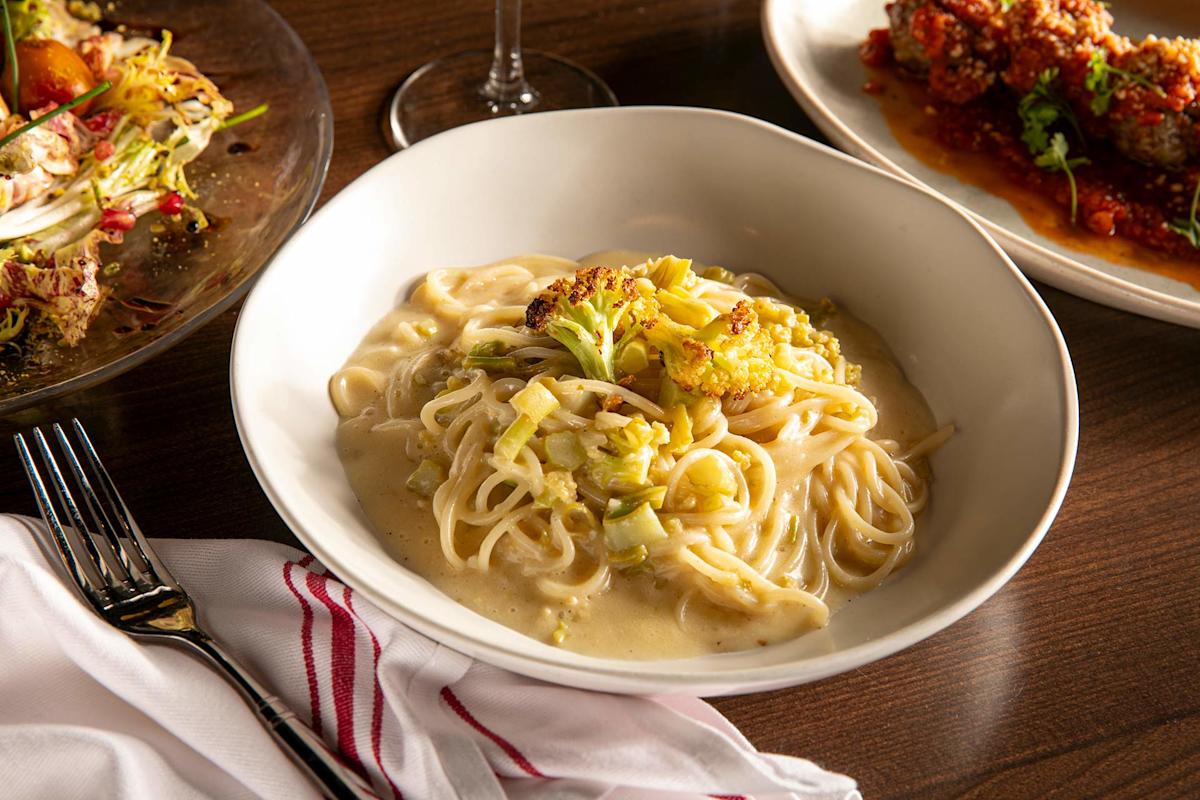 A bowl of creamy pasta topped with roasted cauliflower sits on a wooden table, next to a plate of salad, a dish of meatballs in sauce, a wine glass, a fork, and a white napkin with red stripes.