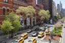 A bustling city street with yellow taxis and cars driving past a historic red-brick building adorned with banners. Trees line the sidewalk, and pedestrians walk along the path. Tall buildings and greenery enhance the urban scene under a clear sky.