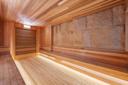 Spacious modern sauna with wooden benches and floor, wood-paneled walls, and a large stone accent wall, all well-lit with natural and built-in lighting.