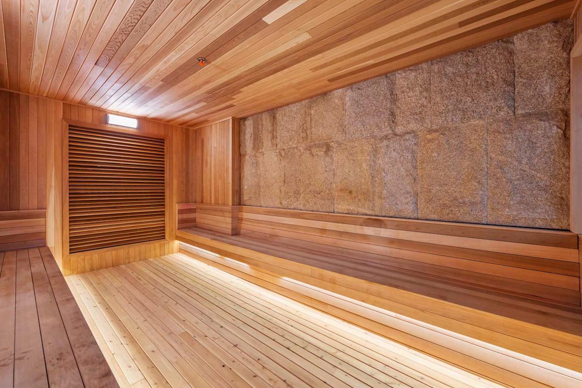 Spacious modern sauna with wooden benches and floor, wood-paneled walls, and a large stone accent wall, all well-lit with natural and built-in lighting.