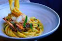 A white bowl filled with bright yellow pasta, garnished with pieces of shrimp, artichoke, and green herbs. The pasta is elegantly twirled. The background is dark, highlighting the vivid colors of the dish.
