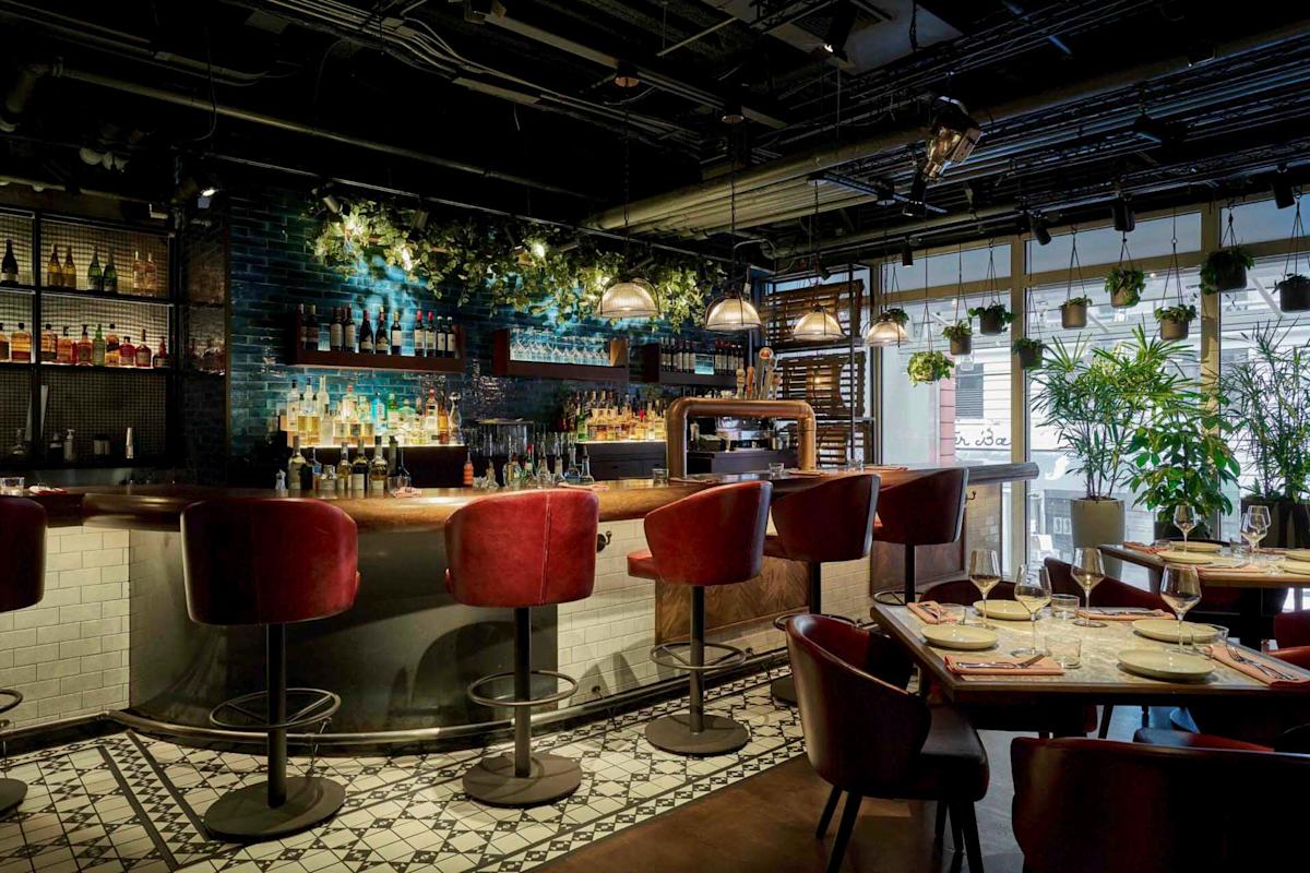 A stylish bar with a curved wooden counter and high red chairs. The back wall features shelves of bottles and hanging pendant lights. The space is decorated with plants, and a table set for dining is visible in the foreground.