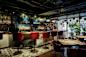 A stylish bar with a curved wooden counter and high red chairs. The back wall features shelves of bottles and hanging pendant lights. The space is decorated with plants, and a table set for dining is visible in the foreground.