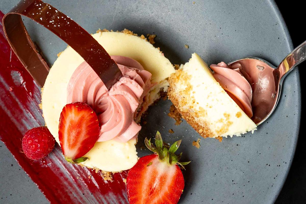 A cheesecake with pink cream, fresh strawberry halves, a raspberry, and a curved chocolate piece sits on a gray plate with a red sauce streak. A spoon holds a bite of cheesecake.