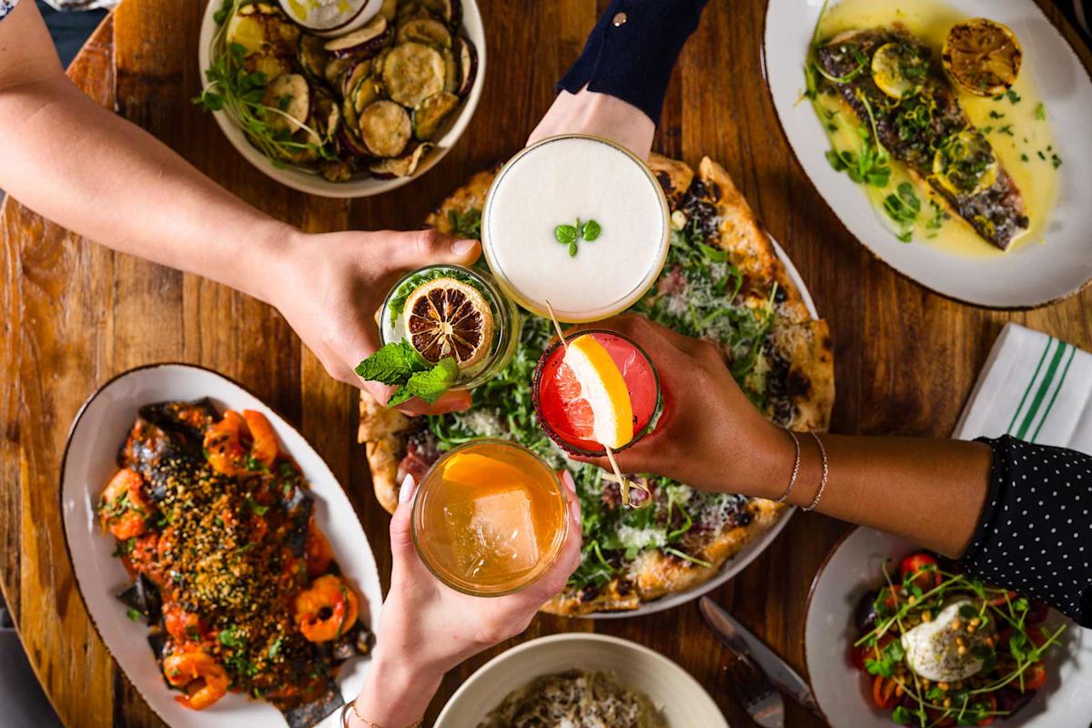 Four people toast with colorful drinks over a wooden table filled with various dishes, including pizza, grilled vegetables, fish, and salads, creating a lively and festive dining atmosphere.
