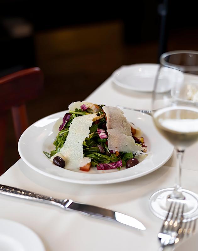 A white plate with mixed greens, olives, tomatoes, and large slices of cheese, set on a white tablecloth beside a wine glass, fork, and knife.