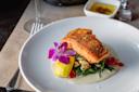 A gourmet dish featuring seared salmon atop a bed of greens and cherry tomatoes, garnished with a purple orchid and lemon wedge. It's presented on a white plate with a glass of wine and a small dish of olive oil in the background.