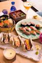A colorful Japanese meal featuring assorted sushi, sashimi, maki rolls, hand rolls, grilled eel over rice, small appetizers, and a cocktail, all beautifully arranged on a table.