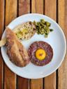 A white plate with beef tartare topped with a raw egg yolk, a slice and a half of crusty bread, and a small pile of chopped pickles, set on a wooden table.