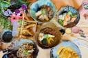A variety of Japanese dishes, including udon, curry rice, sashimi, fried tempura, and katsu, are arranged on a wooden table with a bouquet of flowers and chopsticks beside the plates.