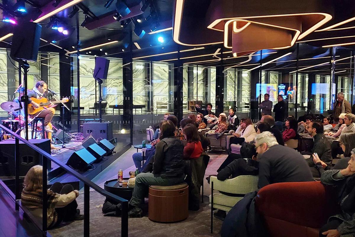 A musician plays guitar and sings on a stage in a modern, stylish venue with glass walls. An audience of people sits on chairs and sofas, watching the performance under dramatic, decorative lighting.