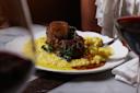 A plate of creamy yellow risotto served with braised meat garnished with herbs, placed on a marble table with glasses of red wine and a person in white seated behind.