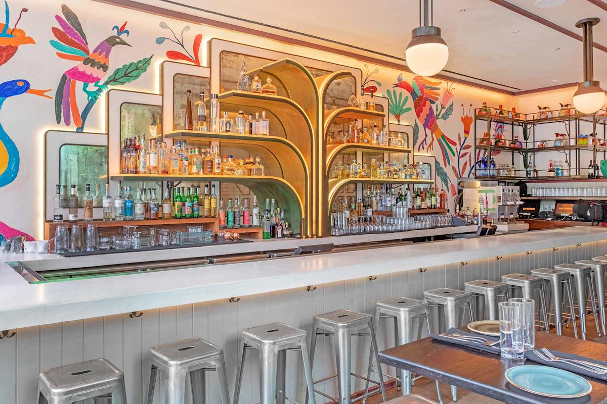 A modern bar with a long white counter, metal stools, and colorful wall art featuring birds and plants. Shelves behind the bar are stocked with bottles, and glassware is neatly arranged. The space is bright and stylish.