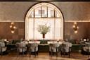 Elegant restaurant interior with a large arched window, textured walls, a long green banquette, neatly set tables, soft lighting, and a tall floral arrangement in the center.