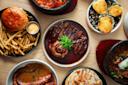An overhead view of various dishes, including steak with sauce and green onions, a burger with fries, baked oysters, grilled octopus, and other assorted foods on a wooden table.