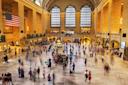 The main hall of Grand Central Terminal in New York City, filled with blurred crowds of people walking. Large arched windows and an American flag are visible. Natural light streams into the spacious interior.