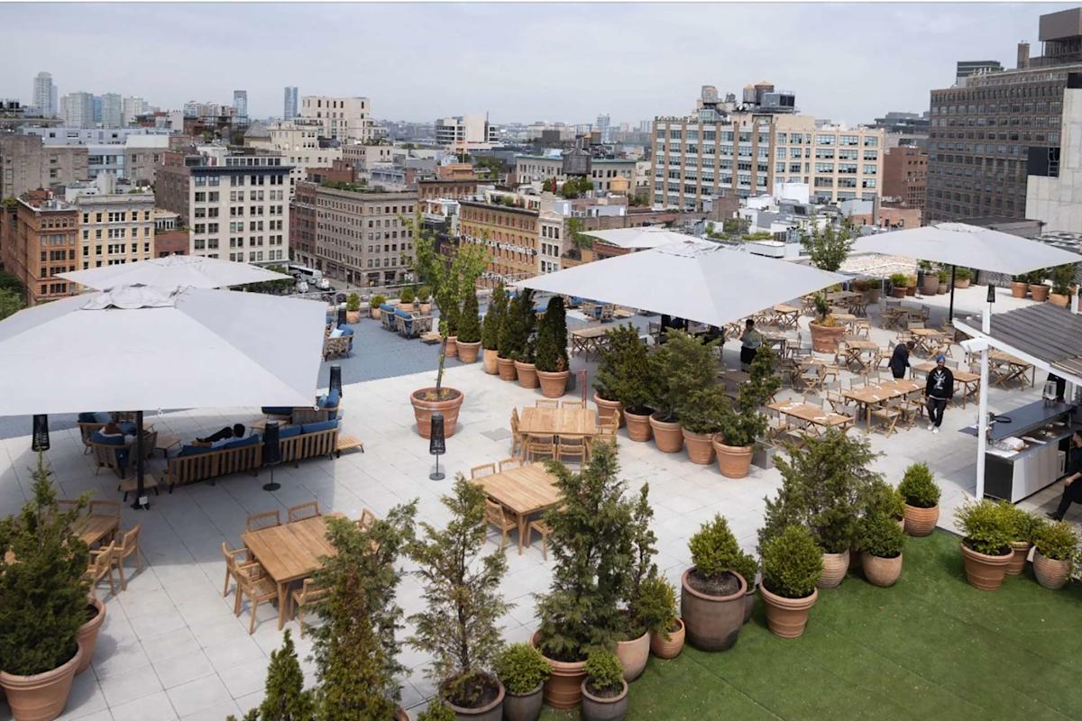 A rooftop terrace with tables, chairs, large umbrellas, and potted plants overlooks a cityscape of mid-rise buildings under a cloudy sky. A few people are seated and walking around the area.