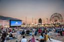 A large crowd sits on blankets and chairs on a beach at sunset, watching a movie on a big outdoor screen. Coney Island rides and bright lights are visible in the background.