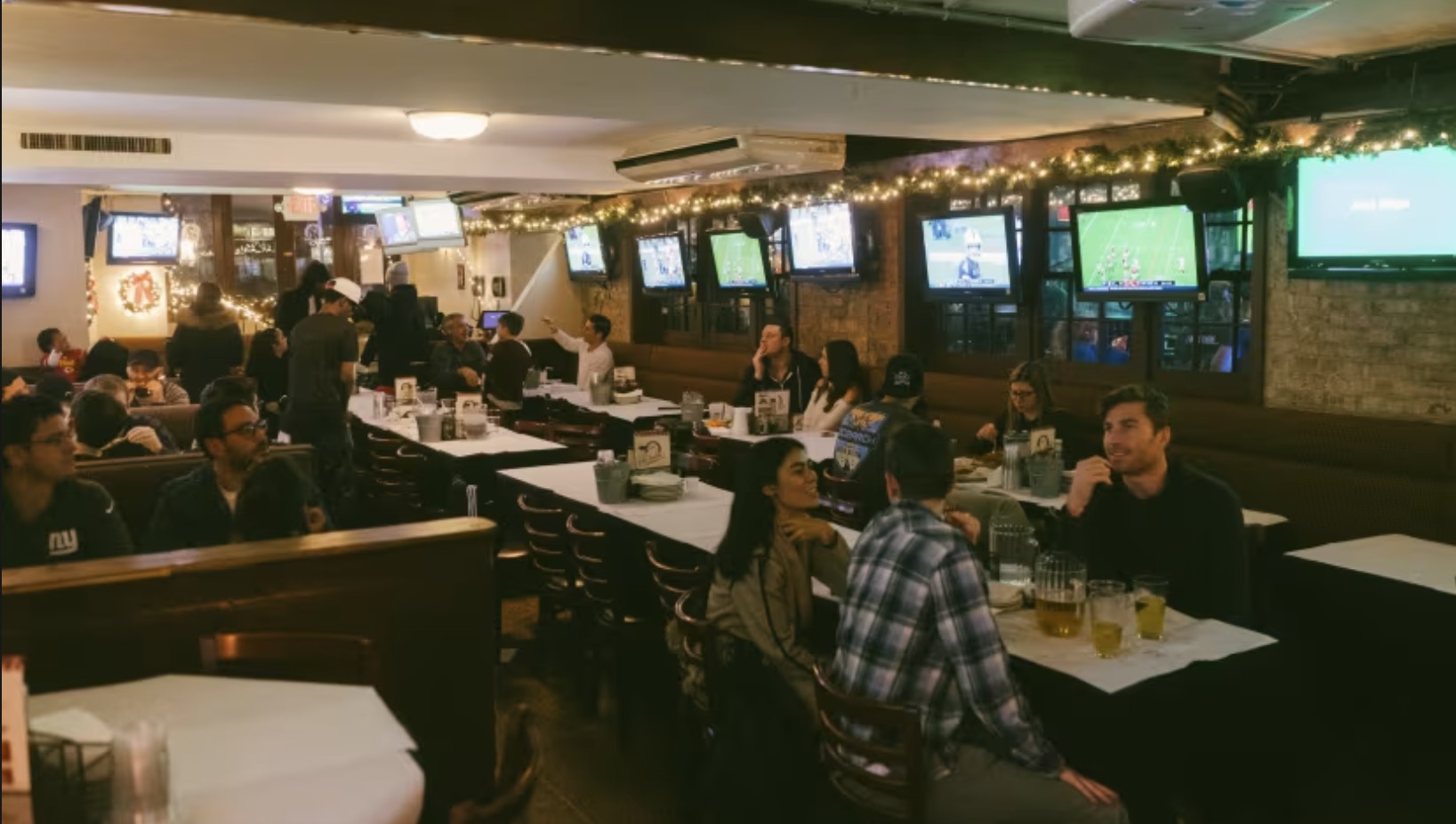 A sports bar with multiple TVs showing games, groups of people sitting at tables with drinks, and string lights decorating the ceiling, creating a lively and casual atmosphere.