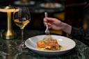 A person holds a fork poised over a dish of grilled salmon with sprouts, served on a white plate. A glass of white wine sits to the side on a dark marbled table, with a small lamp creating a warm ambience.