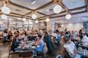 A busy, modern restaurant with people dining at tables, enjoying meals and conversation under round hanging lights. The atmosphere is lively, with staff serving food and a view of the open kitchen in the background.