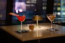 A stylish table setting with two cocktails: a red bird-shaped drink and a classic martini, flanking a bowl of nuts. A small lamp casts a warm glow, and city lights are visible through the window in the background.