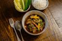 A bowl of spicy stew with vegetables and herbs, served with a side of white rice and fresh greens; a fork and spoon rest beside the dishes on a wooden table.