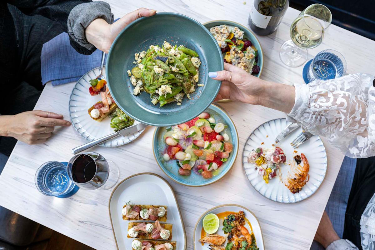 Aerial view of a dining table with multiple plates of colorful dishes, including salads, meat, and appetizers. Two people are holding a green salad bowl, and there are glasses of red and white wine on the table.