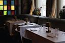 Elegant restaurant interior with tables set for two, each with white tablecloths, glassware, napkins, and small brass lamps; stained glass window and large curtains in the background with floral arrangements.