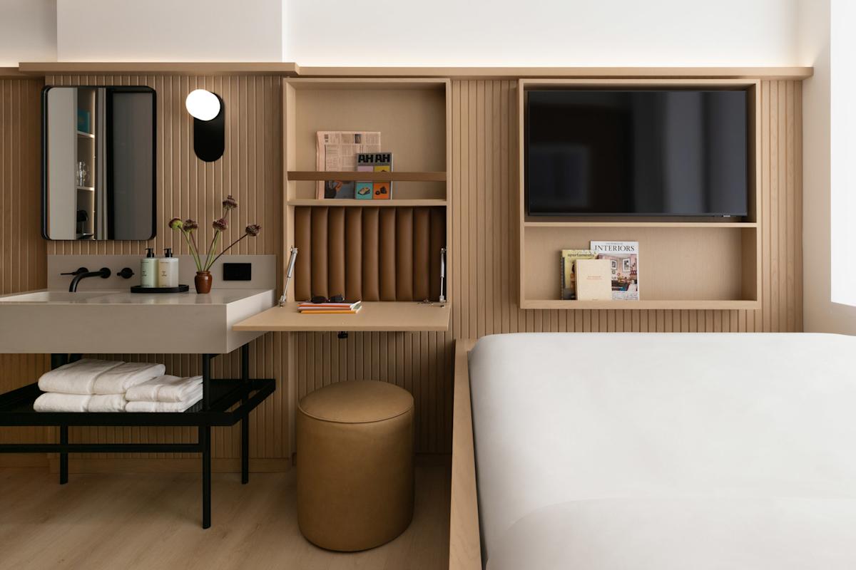 A modern hotel room with a bed, a built-in desk, round stool, wall-mounted TV, open shelves with books, a sink with towels below, and minimal decor in neutral tones.