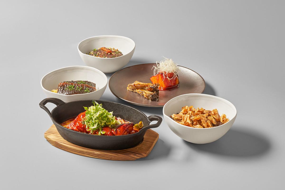Five dishes of gourmet food, including grilled fish, pasta, and vegetables, are elegantly arranged in white bowls and a black cast iron pan on a wooden board against a light grey background.