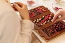 Two people decorate chocolate bars in silicone molds with nuts and dried berries on a table, using their hands to carefully place the toppings. Bowls of toppings are visible in the background.
