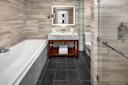 Modern bathroom with a marble bathtub, dark tiled floor, and a glass-enclosed shower. A wooden vanity with a marble countertop holds a mirror with backlighting. Towels are neatly stacked on a shelf under the sink. Walls are tiled in a light pattern.