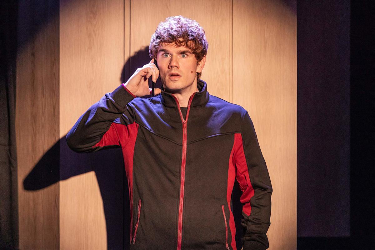A young man with curly hair in a black and red jacket stands under a spotlight, holding a phone to his ear and looking surprised against a wooden background.
