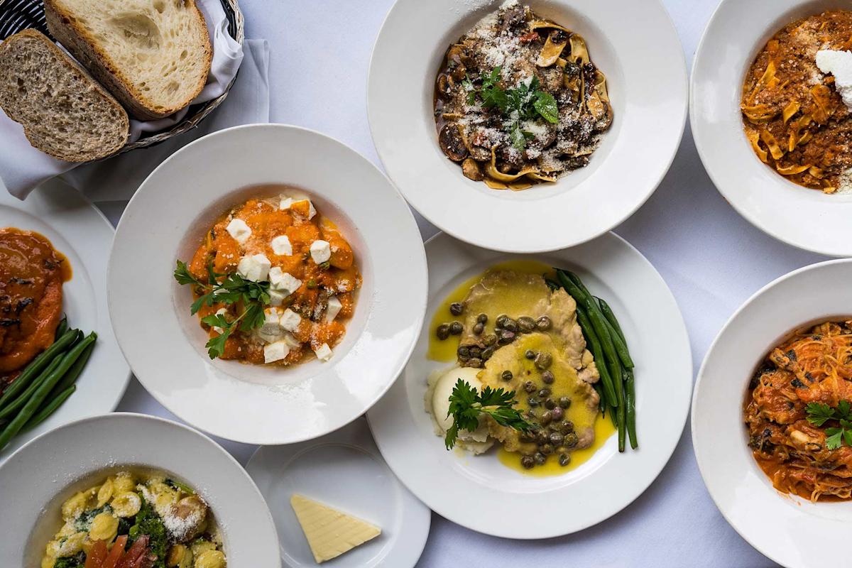 An overhead view of plates with various Italian dishes, including pasta with red and white sauces, ravioli with green beans, gnocchi, bread slices, and a serving of butter, all arranged on a white tablecloth.