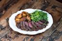 A white plate with sliced steak, roasted potatoes, and a fresh green salad on a rustic wooden table.