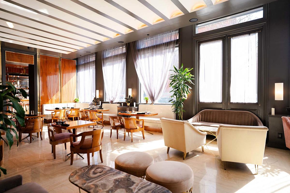 Bright, modern lounge with large windows, sheer curtains, stylish chairs, sofas, and tables. Light floor, dark walls, indoor plants, and soft lighting create a welcoming atmosphere.