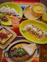 A vibrant Mexican meal with tacos, tortillas, grilled steak with beans, cheesy dip, a dessert with cream, and two colorful drinks, all served on green plates atop a striped tablecloth.