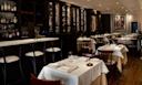 Elegant restaurant interior with white tablecloths, wooden chairs, wine racks on the wall, a stocked bar, and a framed poster. The space is softly lit and neatly arranged for dining.