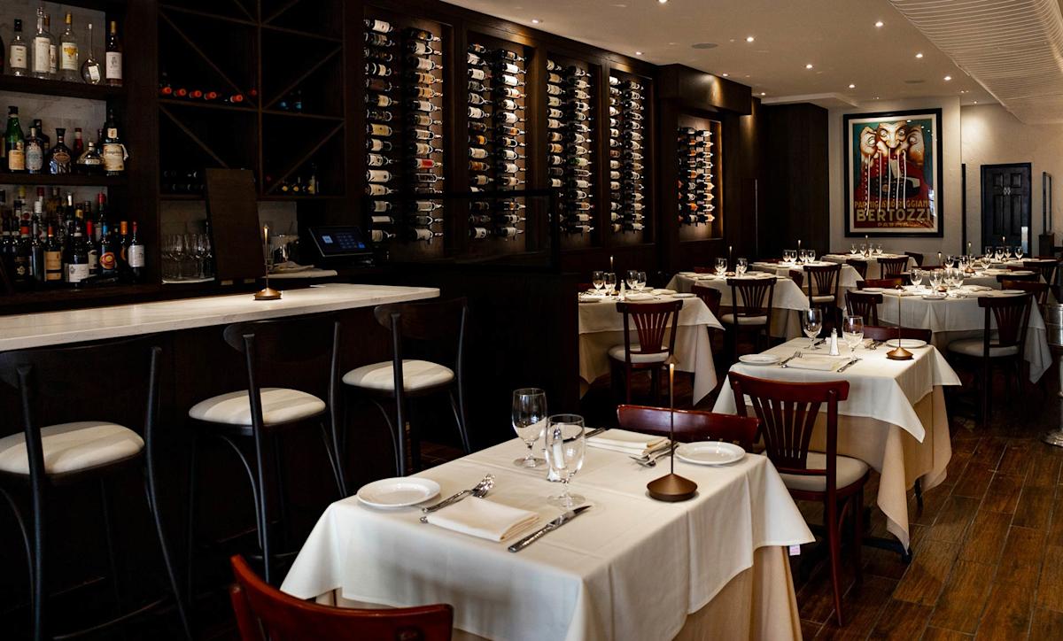 Elegant restaurant interior with white tablecloths, wooden chairs, wine racks on the wall, a stocked bar, and a framed poster. The space is softly lit and neatly arranged for dining.