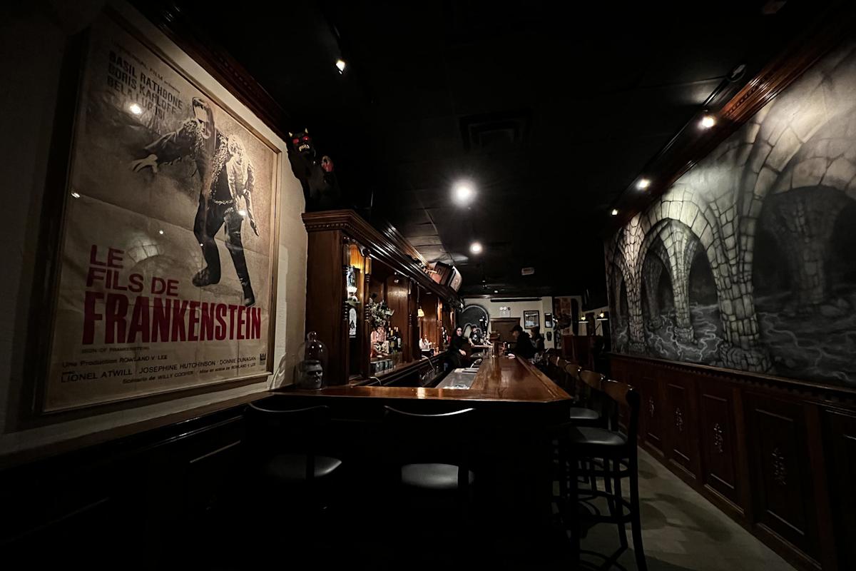 A dimly lit bar with a vintage “Le Fils de Frankenstein” poster on the wall and dark gothic decor, including stone archway artwork and wooden furnishings.