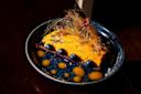 A vibrant dessert on a blue plate, featuring a rectangular chocolate base, bright orange cream, blueberries, yellow sauce dots, gold leaf, spun sugar, and small pink flower petals as garnish.