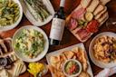 A table set with various dishes, including pasta, salad, grilled asparagus, charcuterie, chips with guacamole, cheese, olives, and a bottle of wine in the center, all arranged on a wooden surface.