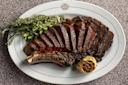 A sliced, grilled bone-in steak is arranged on a white oval plate with a roasted garlic bulb and fresh sprigs of herbs, including rosemary, on the side. The steak is cooked medium-rare and served on a patterned surface.