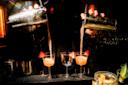 A bartender pours pink cocktails from two shakers into glasses garnished with strawberries on a dimly lit bar counter, with light reflecting off the metal shakers.