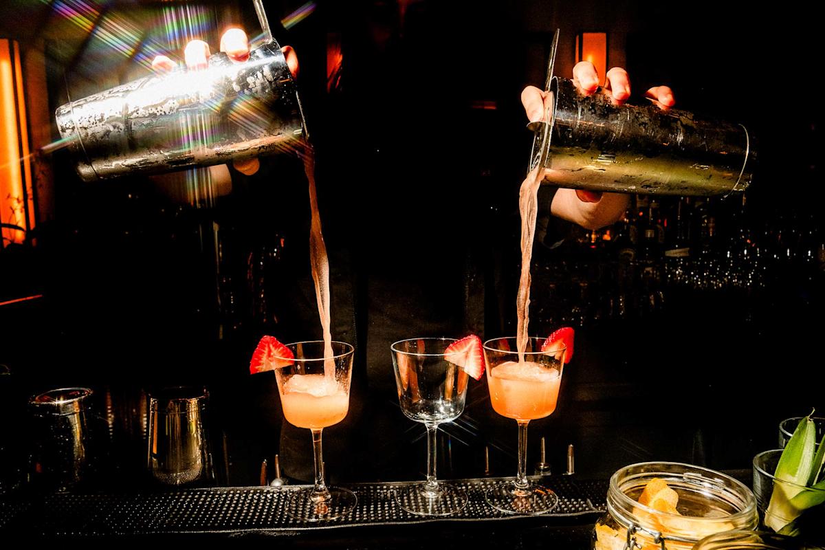 A bartender pours pink cocktails from two shakers into glasses garnished with strawberries on a dimly lit bar counter, with light reflecting off the metal shakers.