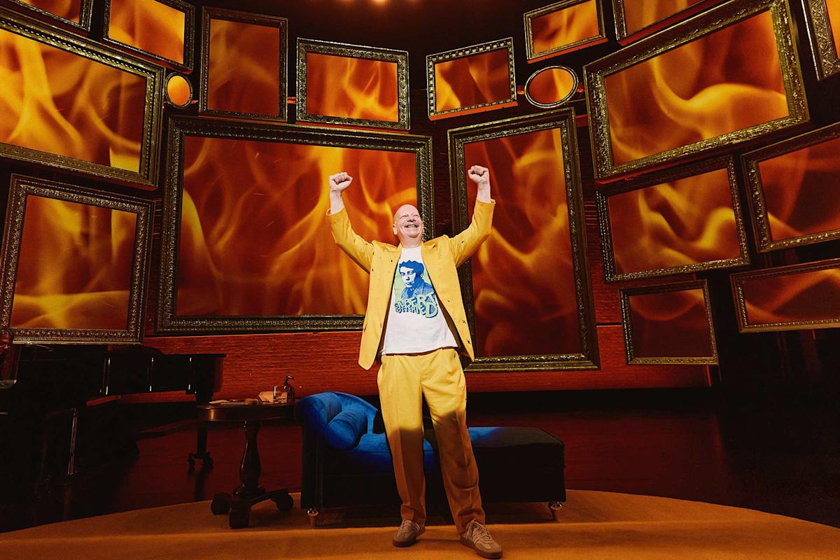 A person in a yellow suit stands center stage with arms raised triumphantly, in front of a backdrop of picture frames filled with images of flames. A blue couch and a piano are visible on stage.