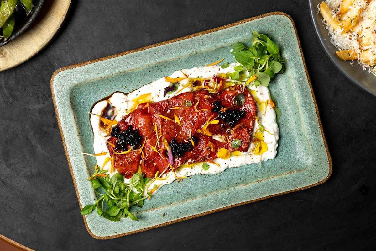 Rectangular plate with roasted red peppers on a bed of creamy white sauce, garnished with herbs, black pearls, microgreens, and edible petals. Some grated cheese and green vegetables are visible on side plates.