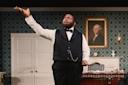 A man in formal attire stands in a vintage room, raising one hand expressively while performing. Behind him is a portrait of George Washington, patterned wallpaper, a desk, and a lit candle.
